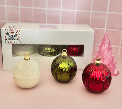 Three glass Christmas ornaments in red, green, and white with a packaging box on a pink tiled background.
